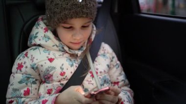 Kışlık şapkalı ve ceketli küçük kız arabada telefonla oynuyor..