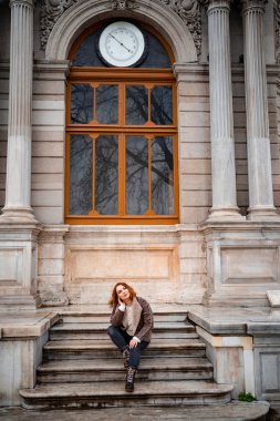 Red haired beautiful girl sitting on the steps of an old building in the rain