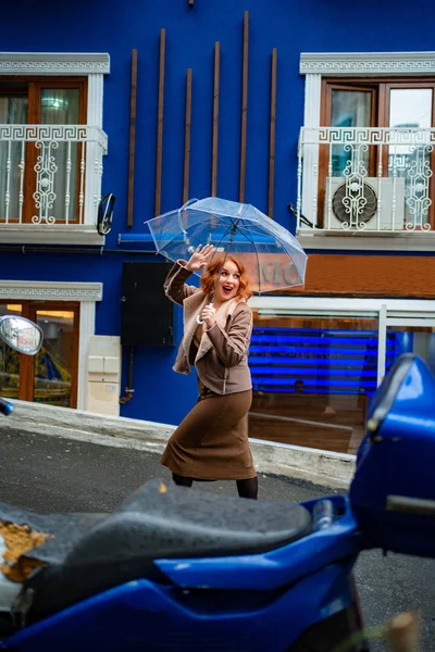Κοκκινομάλλα κορίτσι σε καφέ ρούχα μπλε τοίχο κάτω από ομπρέλα — Φωτογραφία Αρχείου