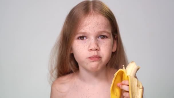 Peu avec des taches de rousseur mange une banane, regardant caméra — Video