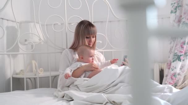 Мама в ліжку з дитиною на руках дивиться на телефон — стокове відео