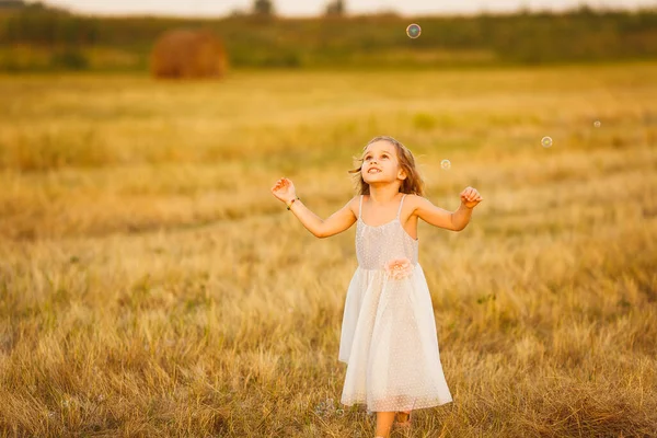 Menina perseguindo bolhas de sabão no campo por do sol — Fotografia de Stock