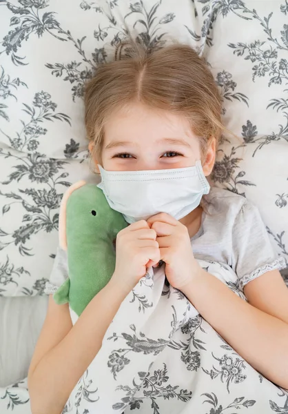 Girl lying in bed in medical mask. dinosaur — Stock Photo, Image