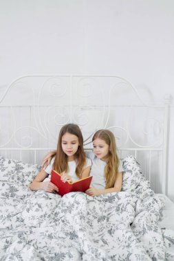 Kız kardeşler birlikte kitap okuyor, yatakta oturuyorlar.