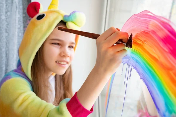 Bürste. Teenie-Mädchen im Kigurumi zeichnet Regenbogenfenster — Stockfoto