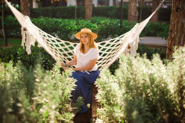 Şapkalı ve mavi etekli kadın Park 'ta hamakta oturuyor.
