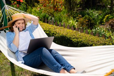 Şapkalı kadın hamakta yatıyor, telefona cevap veriyor.
