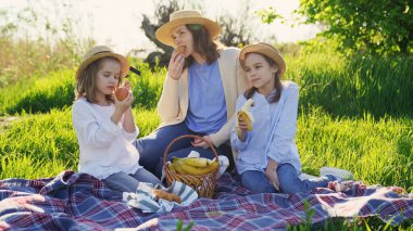 Anneler ve kızları çimenlikte piknikte ve meyve yiyorlar.