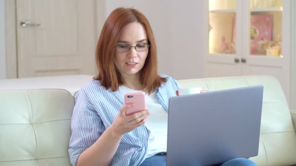 Freelancer woman working at computer, drinks tea and writes message to phone. — Stock Video