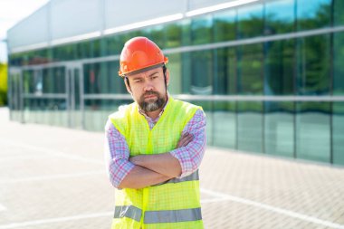 Yelek ve koruyucu miğfer giyen ustabaşı yeni yapının arka planında duruyor..