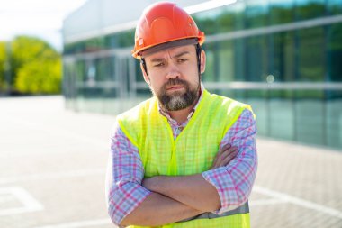 Yelek ve koruyucu miğfer giyen ustabaşı yeni yapının arka planında duruyor..