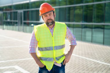 Yelek ve koruyucu miğfer giyen ustabaşı yeni yapının arka planında duruyor..