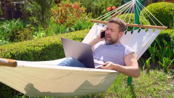 Uomo si trova in amaca e di lavoro sul computer, giurando al telefono — Video Stock