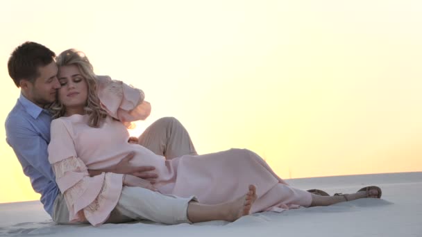 A woman lays on a mans chest as they sit on the beach and watch the sun set together — Stock Video