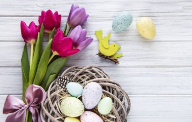 Yuvasında renkli yumurtalar ve beyaz ahşap arka planda pembe laleler olan Paskalya arkaplanı. Kopyalama alanı olan üst görünüm