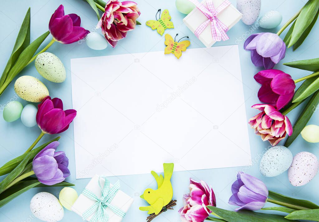 Greeting card. Spring tulip flowers, easter eggs and  gift box  on pastel blue background from above in flat lay style.