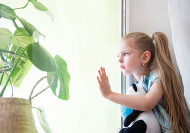 Evdeki pencerenin yanında üzgün bir kız çocuğu. Karantinaya alın. Koronavirüs salgını. Evde kal konsepti.
