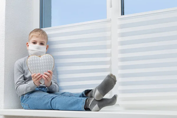 Üzgün çocuk, koruyucu maskeli ve beyaz kalpli pencere eşiğinde oturuyor. — Stok fotoğraf