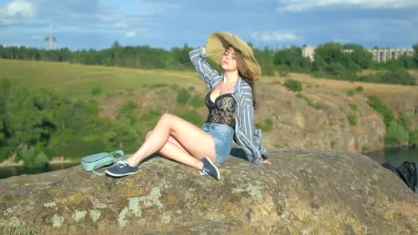 Молода дівчина в капелюсі сидить на скелі — стокове відео