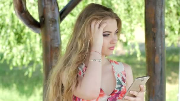 Retrato de una joven relajada en un parque de verano leyendo un mensaje de texto en su teléfono móvil . — Vídeo de stock