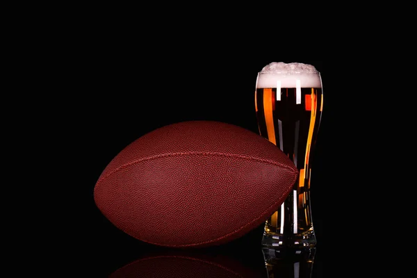 Beer glass with dark beer foam and American football ball on black background.