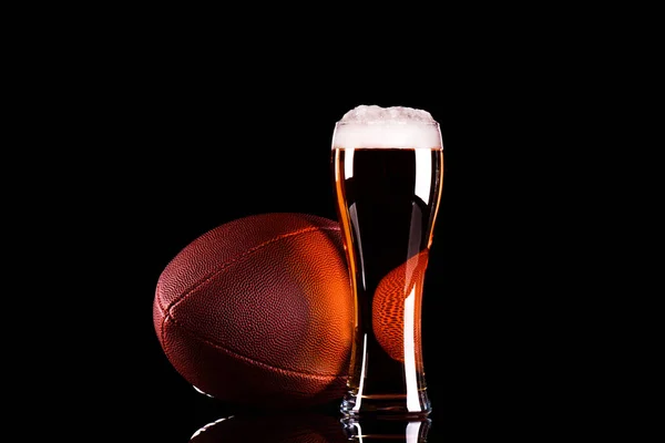 Copo de cerveja com espuma de cerveja escura e bola de futebol americano no fundo preto — Fotografia de Stock