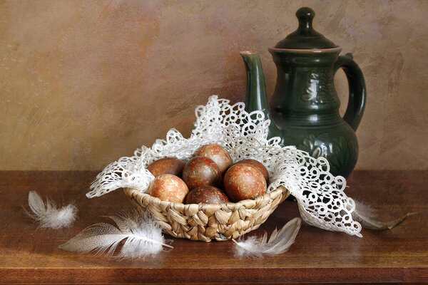 Chicken egg on an openwork napkin, ceramic teapot, feathers . Still life on a dark old background .