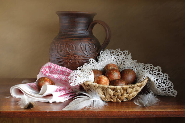 Chicken egg on an openwork napkin, clay jug, feathers . Still life on a dark old background .