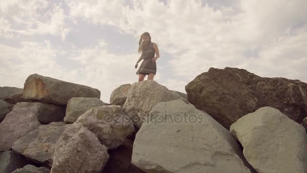 Дівчина йде вздовж скелястого берега моря, балансуючи на вузькому хребті, тримаючи в руці мобільний телефон. Повільний рух — стокове відео