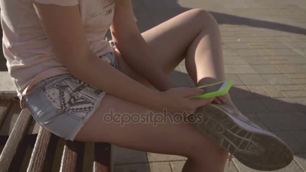 Uma menina senta-se em um banco e segura um smartphone em sua mão — Vídeo de Stock