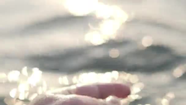 Male hand playfully and gently touches the surface of the sea in the glare of the sun at sunset, close-up, — ストック動画