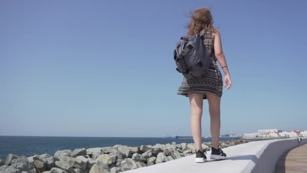 Una ragazza con uno zaino e cuffie wireless cammina lungo la riva del mare in una giornata di sole . — Video Stock