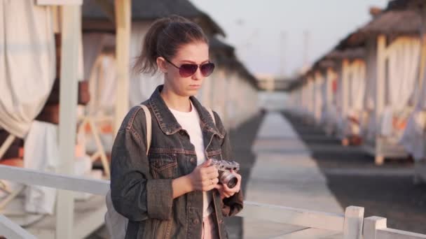 Chica en la playa con una cámara fotográfica la puesta del sol. Tumbonas vacías — Vídeos de Stock