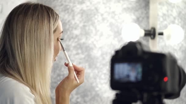 De mooie blonde make-up kunstenaar is het aanbrengen van oogschaduw kijken in een spiegel — Stockvideo