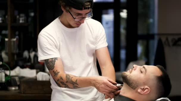 Barber in black cap clipping clients beard in barbershop. — 비디오