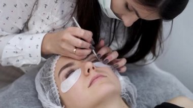 Tek kullanımlık maskeli esmer bir Avrupalı kirpik sanatçısı kirpiklerin kaplamasını yapıyor.
