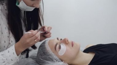 Tek kullanımlık maskeli esmer bir Avrupalı kirpik sanatçısı kirpiklerin kaplamasını yapıyor.