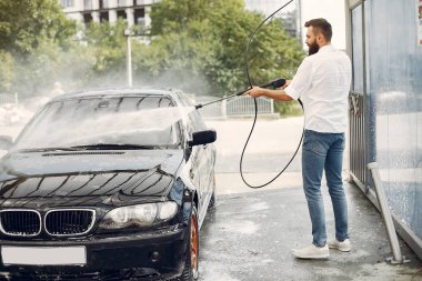 Beyaz gömlekli bir adam arabasını yıkıyor.