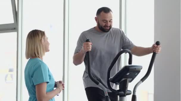 Пациент делает упражнения на велосипеде в тренажерном зале с терапевтом — стоковое видео