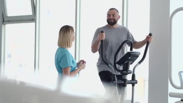 Patienten tränar på spinncykel i gymmet med terapeut — Stockvideo
