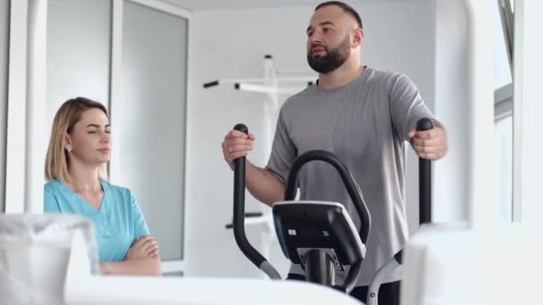 Pacjent ćwiczy na rowerze spinningowym w siłowni z terapeutą — Wideo stockowe