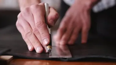 The tanner is cutting black leather sheet with a razor and metre