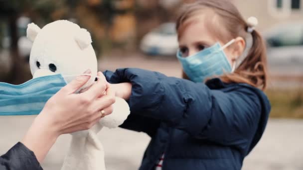 Uma menina em uma máscara com seu ursinho de pelúcia usando uma máscara descartável — Vídeo de Stock