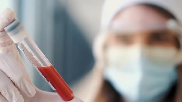 Mulher doutor em um chapéu médico e uma máscara está segurando um frasco com sangue — Vídeo de Stock
