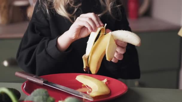 Das kleine Mädchen schält und schneidet in einer modernen Küche eine Banane — Stockvideo