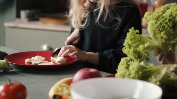 Das kleine Mädchen schält und schneidet in einer modernen Küche eine Banane — Stockvideo