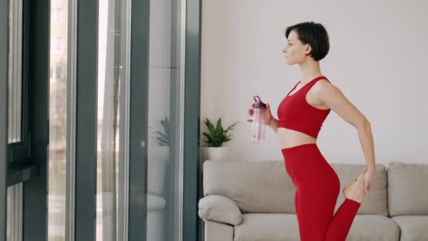 Фит-женщина в красном костюме растягивает ноги, стоя с фитнес-бутылкой. — стоковое видео