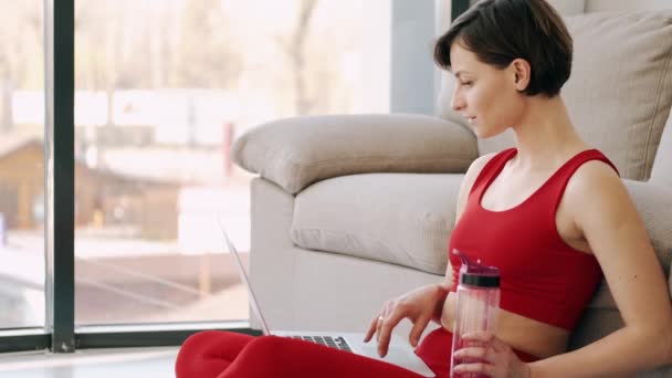 Mujer deportiva con una botella de fitness está trabajando con un ordenador portátil en una habitación moderna — Vídeo de stock