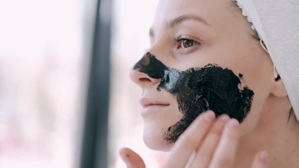 Jolie femme avec des cheveux enveloppés dans une serviette applique un masque noir sur le visage — Video