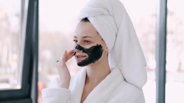 Jolie femme avec des cheveux enveloppés dans une serviette applique un masque noir sur le visage — Video